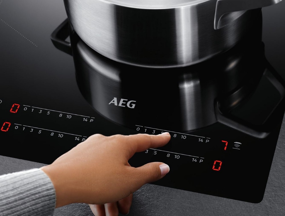 A hand operating the touch control panel of the AEG IKE64441XB induction hob to adjust the temperature for a pot.