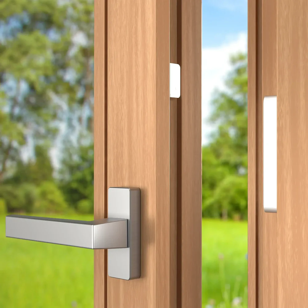 The magnetic window sensor (small and large part) is discreetly mounted on an open wooden window frame, with a green garden in the background.