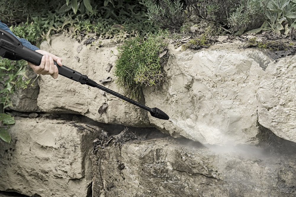 A hand holding the spray lance of the Kärcher pressure washer, cleaning a rough, mossy stone wall in the garden from dirt.
