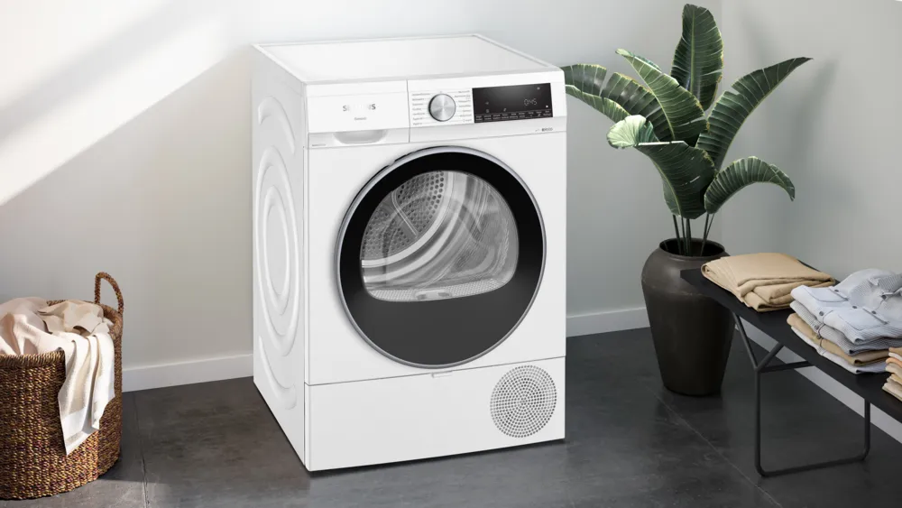 The white Siemens dryer placed in a modern laundry space with a large potted plant, laundry basket, and stacked laundry.